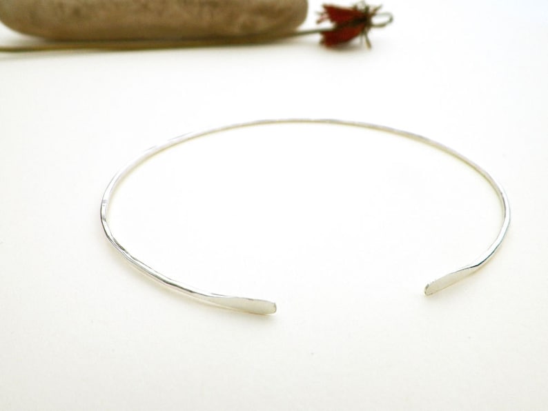 May include: A simple silver choker necklace with a hammered finish. The necklace is open in the back and has a slightly curved shape.