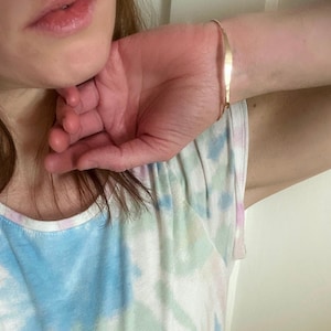 K&ouml;nnte beinhalten: Ein zartes, goldfarbenes Armband. Das Armband ist d&uuml;nn und hat ein schlichtes, elegantes Design. Das Armband wird am Handgelenk getragen. Die Person tr&auml;gt ein Tanktop mit Batikmuster.