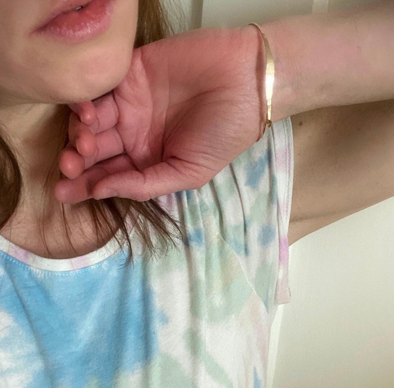 K&ouml;nnte beinhalten: Ein zartes, goldfarbenes Armband. Das Armband ist d&uuml;nn und hat ein schlichtes, elegantes Design. Das Armband wird am Handgelenk getragen. Die Person tr&auml;gt ein Tanktop mit Batikmuster.