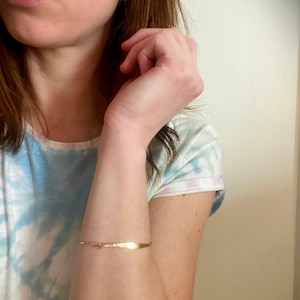 K&ouml;nnte beinhalten: Ein zartes, goldfarbenes Armband mit geh&auml;mmerten Textur. Das Armband ist d&uuml;nn und umschlie&szlig;t das Handgelenk. Das Design weist ein kleines, dekoratives Element in der N&auml;he des Verschlusses auf. Das Armband hat einen Umfang von etwa 15 cm.