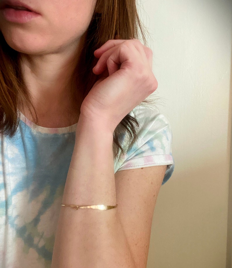 K&ouml;nnte beinhalten: Ein zartes, goldfarbenes Armband mit geh&auml;mmerten Textur. Das Armband ist d&uuml;nn und umschlie&szlig;t das Handgelenk. Das Design weist ein kleines, dekoratives Element in der N&auml;he des Verschlusses auf. Das Armband hat einen Umfang von etwa 15 cm.