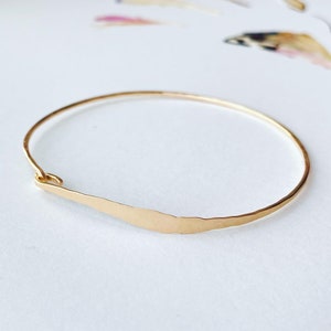 K&ouml;nnte beinhalten: Ein goldfarbenes Armreif-Armband mit geh&auml;mmertem, strukturiertem Finish. Das Armband hat ein schlichtes, elegantes Design mit einem geschwungenen Barren-Detail. Das Armband wird auf einer wei&szlig;en Oberfl&auml;che gezeigt.