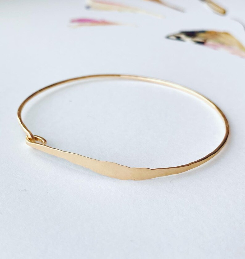 K&ouml;nnte beinhalten: Ein goldfarbenes Armreif-Armband mit geh&auml;mmertem, strukturiertem Finish. Das Armband hat ein schlichtes, elegantes Design mit einem geschwungenen Barren-Detail. Das Armband wird auf einer wei&szlig;en Oberfl&auml;che gezeigt.