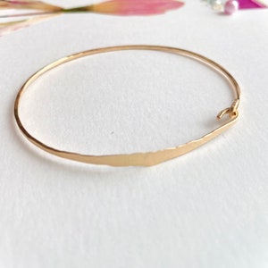 K&ouml;nnte beinhalten: Ein goldfarbenes Armreif-Armband mit geh&auml;mmerten Texturen. Das Armband hat ein schlichtes, elegantes Design mit einem Hakenverschluss. Der Hintergrund ist wei&szlig; mit weichen floralen Akzenten.