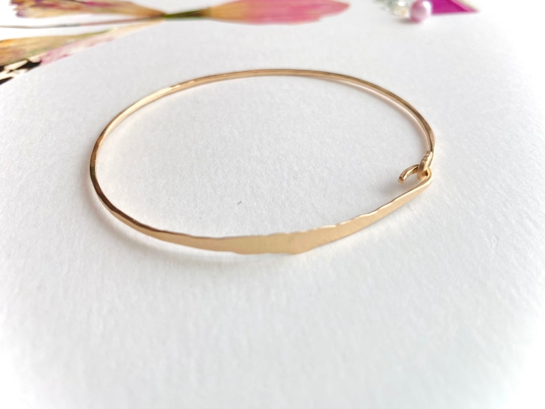 K&ouml;nnte beinhalten: Ein goldfarbenes Armreif-Armband mit geh&auml;mmerten Texturen. Das Armband hat ein schlichtes, elegantes Design mit einem Hakenverschluss. Der Hintergrund ist wei&szlig; mit weichen floralen Akzenten.