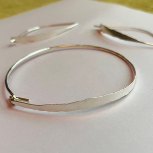 K&ouml;nnte beinhalten: Ein silbernes Armreif-Armband mit geh&auml;mmertem Muster und zwei passenden ohrringf&ouml;rmigen Bl&auml;ttern. Das Armband hat einen einfachen Hakenverschluss. Der Schmuck wird auf einer hellrosa Oberfl&auml;che pr&auml;sentiert.