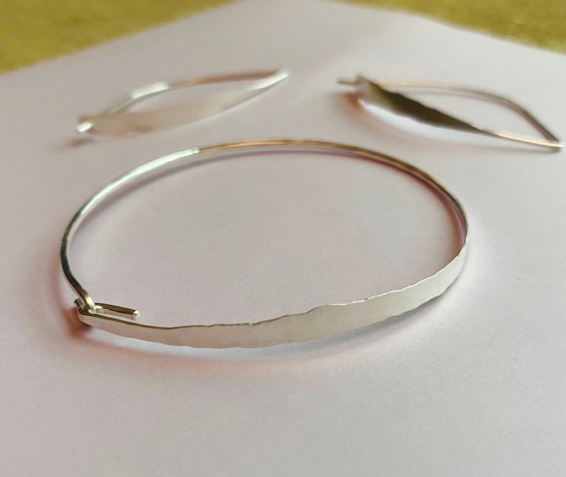 K&ouml;nnte beinhalten: Ein silbernes Armreif-Armband mit geh&auml;mmertem Muster und zwei passenden ohrringf&ouml;rmigen Bl&auml;ttern. Das Armband hat einen einfachen Hakenverschluss. Der Schmuck wird auf einer hellrosa Oberfl&auml;che pr&auml;sentiert.