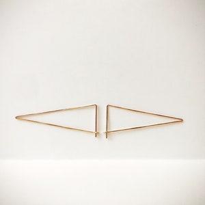 May include: Two gold-colored, geometric earrings. Each earring is a triangle shape, with a straight base and two angled sides. The earrings are displayed on a white background.