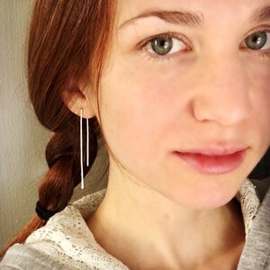May include: A woman with long, red hair wearing a pair of long, thin, silver earrings. The earrings are straight and dangle down from her earlobes.