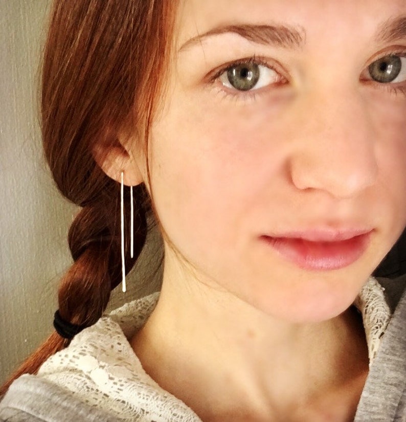 May include: A woman with long, red hair wearing a pair of long, thin, silver earrings. The earrings are straight and dangle down from her earlobes.