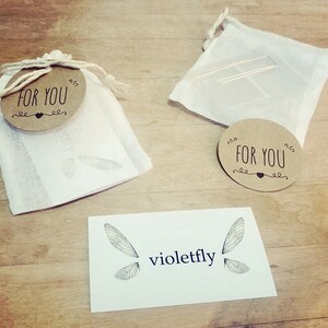 Puede incluir: Dos peque&ntilde;as bolsas de tela blanca con etiquetas circulares marrones que dicen "Para ti" con un s&iacute;mbolo de coraz&oacute;n. Las bolsas est&aacute;n sobre una superficie de madera con una tarjeta blanca que dice "violetfly" y tiene un dibujo de dos alas de lib&eacute;lula.