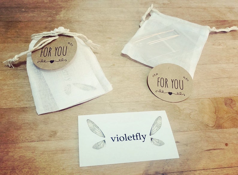 Puede incluir: Dos peque&ntilde;as bolsas de tela blanca con etiquetas circulares marrones que dicen "Para ti" con un s&iacute;mbolo de coraz&oacute;n. Las bolsas est&aacute;n sobre una superficie de madera con una tarjeta blanca que dice "violetfly" y tiene un dibujo de dos alas de lib&eacute;lula.