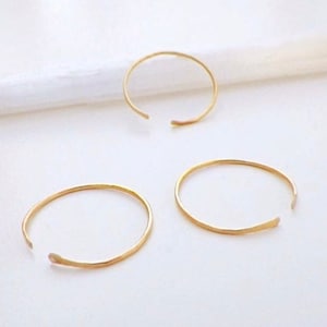 Puede incluir: Tres anillos de aro abiertos de color dorado sobre una superficie blanca. Los anillos son simples y delicados, con una textura martillada.