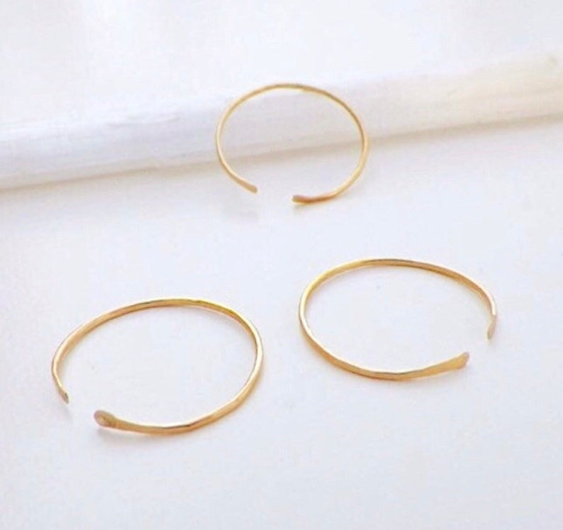 Puede incluir: Tres anillos de aro abiertos de color dorado sobre una superficie blanca. Los anillos son simples y delicados, con una textura martillada.