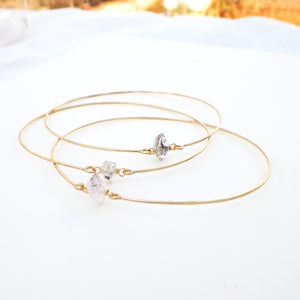 May include: Three delicate gold-colored bangles, each adorned with a clear, irregularly shaped crystal. The crystals are attached to the bangles with small gold-colored connectors. The bangles are displayed on a white surface.