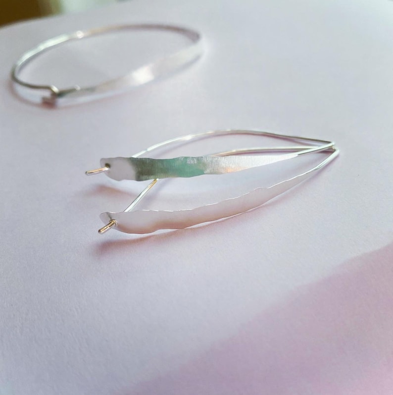 K&ouml;nnte beinhalten: Zwei Schmuckst&uuml;cke aus Silber. Eines ist ein Armband, das andere ein Paar Ohrringe. Die Ohrringe haben ein einzigartiges Design mit einer langen, strukturierten, blattartigen Form. Das Armband ist ein einfaches, d&uuml;nnes Band. Beide Teile haben ein gl&auml;nzendes, metallisches Finish.