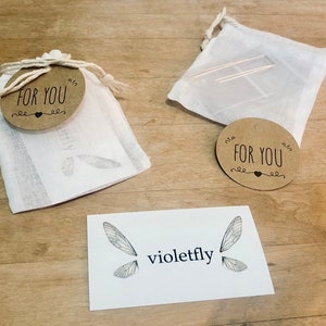 May include: Two small white muslin bags with brown paper tags that say "For You" and a small heart. The bags are on a wooden surface with a white card that says "violetfly" and has a drawing of a dragonfly on it.
