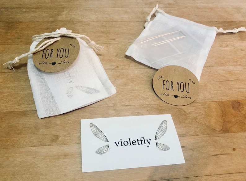 May include: Two small white muslin bags with brown paper tags that say "For You" and a small heart. The bags are on a wooden surface with a white card that says "violetfly" and has a drawing of a dragonfly on it.