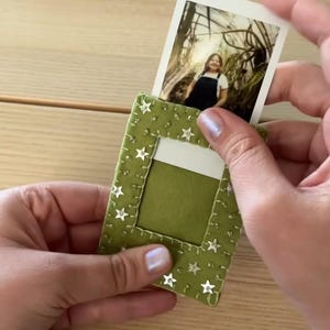 May include: A rectangular olive green felt photo holder featuring a stitched border and small silver star-shaped embellishments. The holder has a central cutout window designed to display a small rectangular photograph of a person.