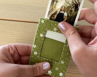 May include: A rectangular olive green felt photo holder featuring a stitched border and small silver star-shaped embellishments. The holder has a central cutout window designed to display a small rectangular photograph of a person.