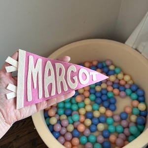 May include: A pink felt pennant with the name "MARGOT" in white block letters. The pennant is held in front of a beige plastic tub filled with colorful plastic balls in shades of blue, green, peach, and yellow.