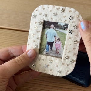 Peut inclure: Un cadre photo rectangulaire en feutrine blanc cassé aux coins arrondis, orné d'une bordure de petites paillettes argentées en forme d'étoile et de perles transparentes. Le cadre contient une photo d'un adulte et d'un enfant marchant dehors.