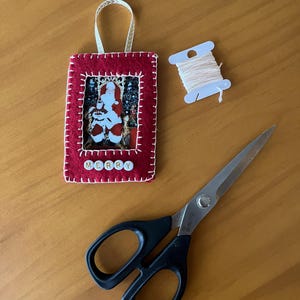 May include: A red felt Christmas ornament with a white stitched border. The ornament has a picture frame with a photo of Santa Claus inside. The word "MERRY" is spelled out with white letter beads on the bottom of the ornament. The ornament is hanging from a white ribbon. There is also a spool of white thread and a pair of black handled scissors on a wooden surface.