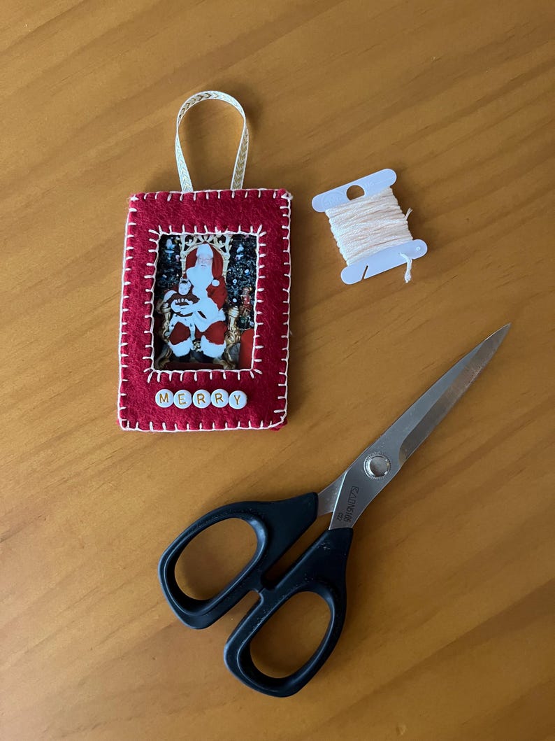 May include: A red felt Christmas ornament with a white stitched border. The ornament has a picture frame with a photo of Santa Claus inside. The word "MERRY" is spelled out with white letter beads on the bottom of the ornament. The ornament is hanging from a white ribbon. There is also a spool of white thread and a pair of black handled scissors on a wooden surface.