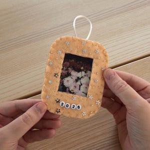 May include: A rectangular peach-colored felt ornament with rounded corners, featuring a central photo of pink and white flowers. The frame is decorated with small silver star sequins and white beads spelling 2026 at the bottom.