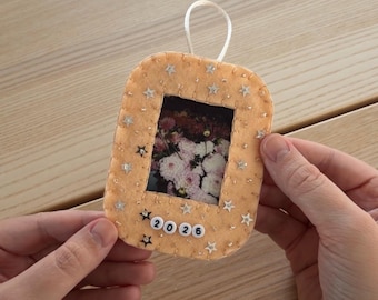 May include: A rectangular peach-colored felt ornament with rounded corners, featuring a central photo of pink and white flowers. The frame is decorated with small silver star sequins and white beads spelling 2026 at the bottom.