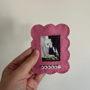 Puede incluir: Un marco de fotos de fieltro rosa con un borde festoneado. El marco tiene una foto de una familia de tres personas dentro y el nombre "MARGOT" escrito en cuentas blancas debajo de la foto.