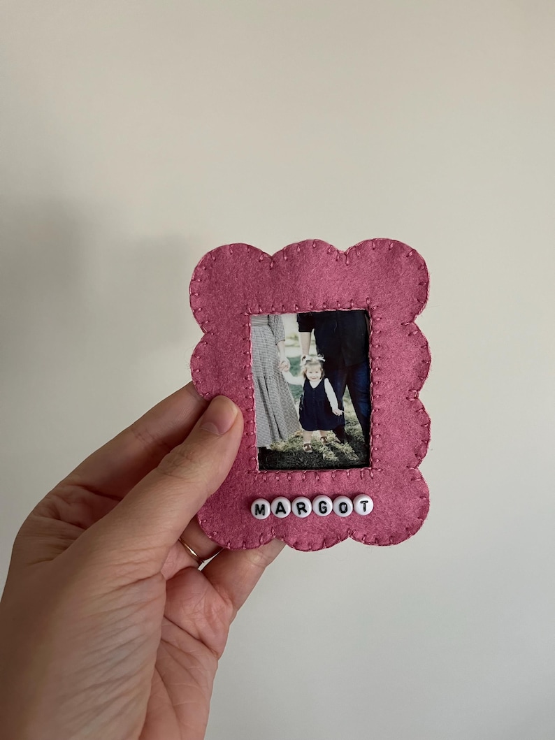 May include: A pink felt picture frame with a scalloped edge. The frame has a photo of a family of three inside and the name "MARGOT" spelled out in white beads below the photo.