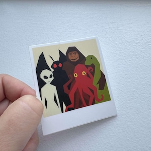 Peut inclure: Un autocollant carré représentant un groupe de cryptides stylisés. Les créatures comprennent un extraterrestre blanc, un homme-papillon noir, un bigfoot brun, une créature rouge en forme de poulpe et une figure verte en forme de dinosaure. L'autocollant a une bordure blanche et un fond crème.
