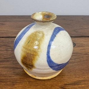 May include: A small, spherical ceramic vase with a cream-colored base glaze. The surface features hand-painted abstract markings in deep blue and earthy brown tones. It has a narrow, flared neck opening and a smooth, rounded body shape.