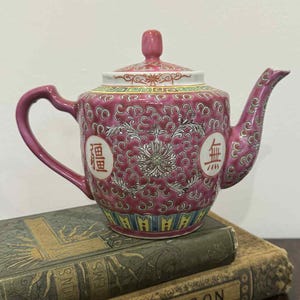 May include: A pink ceramic teapot with a floral pattern and Chinese characters. The teapot sits atop two antique books with gold lettering. The teapot has a curved handle and spout, and a matching lid.