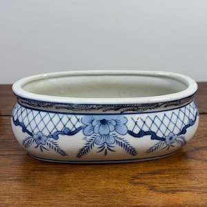 May include: An oval ceramic planter featuring a white base with blue floral patterns and a lattice design around the rim. The bowl has a smooth, glazed finish with a wide opening and a rounded body, showcasing intricate hand-painted blue details.