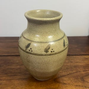 May include: A small, speckled beige ceramic vase with a flared rim and a rounded body. The vase features a horizontal band around the center decorated with a repeating pattern of dark brown leaf-like shapes and small dots.