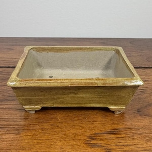May include: A rectangular ceramic bonsai pot with a glazed mustard-yellow exterior and a light beige interior. The vessel features a wide rim, slightly tapered sides, and four small feet at the base, designed for plant cultivation.