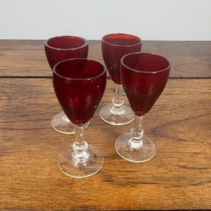 May include: A set of four small stemmed glasses featuring deep red bowls and clear glass stems and circular bases. Each glass has a rounded cup shape and a slender stem with a small decorative knob detail near the base of the bowl.
