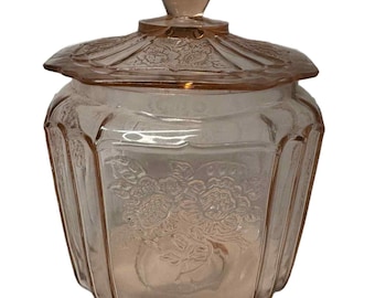 Pode incluir: Um frasco de vidro rosa com tampa. O frasco tem formato hexagonal com desenhos florais gravados no vidro. A tampa tem uma borda decorativa e um pequeno botão na parte superior. O frasco é provavelmente vintage ou antigo.