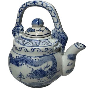 Könnte beinhalten: Weiß-blaue Keramik-Teekanne mit Drachendesign. Die Teekanne hat einen gebogenen Griff, einen Ausguss und einen Deckel mit einem kleinen Knopf. Der Korpus ist mit einem blauen Drachen und traditionellen chinesischen Mustern verziert.