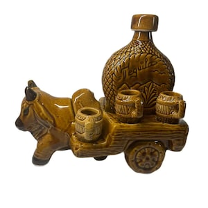 May include: A brown ceramic tequila bottle and shot glass set shaped like a bull pulling a cart. The bottle is textured and labeled "Tequila". The cart holds three shot glasses. The set is a decorative item.