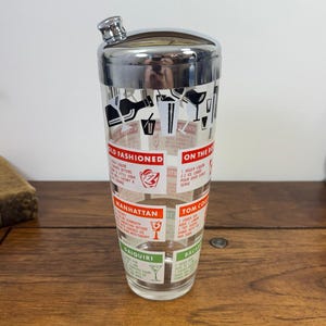 May include: A clear glass cocktail shaker with a polished chrome lid featuring a built-in strainer. The exterior is printed with black, red, and green graphics of various cocktail recipes including Old Fashioned, Manhattan, and Daiquiri.