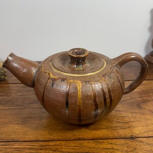 May include: A brown ceramic teapot with a rounded body, a curved handle, and a short spout. The surface features vertical ridges and a glossy glaze with dark brown drip patterns running down the sides from the lid and rim area.