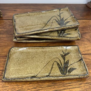 May include: A set of four rectangular ceramic plates with a speckled tan glaze and a dark brown hand-painted bamboo motif. The plates feature slightly raised edges and a glossy finish, with one plate placed in the foreground and three stacked behind.
