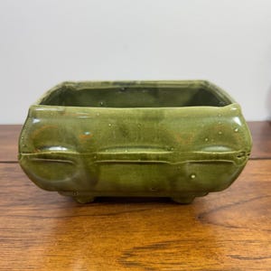 May include: A square, olive green ceramic planter with a glossy finish. The exterior features a horizontal raised ridge detail along the center and rounded corners. The vessel is supported by four small, integrated feet at the base.