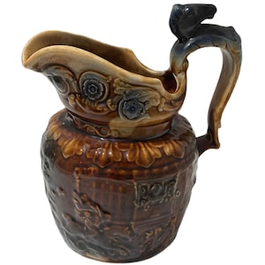 Könnte beinhalten: Ein brauner Keramikkrug mit dekorativem Griff und Ausguss. Der Krug weist florale und reliefartige Designs auf, mit einem dunkelblauen Akzent am Griff. Der Krug hat ein Fassungsvermögen von etwa 1 Liter.