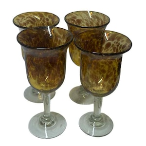 Puede incluir: Cuatro copas de vino color ámbar con un patrón moteado. Las copas tienen forma de cuenco redondeado y se apoyan sobre tallos y bases transparentes. Las copas están dispuestas en grupo sobre un fondo blanco.