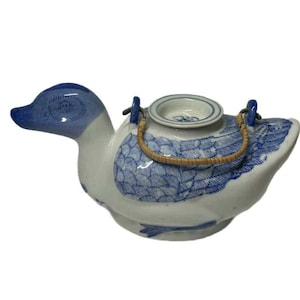 May include: A ceramic duck-shaped teapot in white and blue. The teapot features a blue head and tail, with blue patterned wings and body. It has a woven handle and a small opening on top.