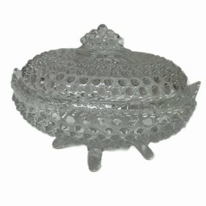 May include: A clear, glass candy dish with a lid. The dish has a textured, hobnail pattern and sits on three small feet. The lid is topped with a decorative finial. The dish is designed to hold small candies or other treats.